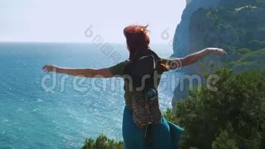 一个年轻的红发女孩旅行者戴着牛仔帽和背包<strong>站在山顶</strong>上，双臂张开